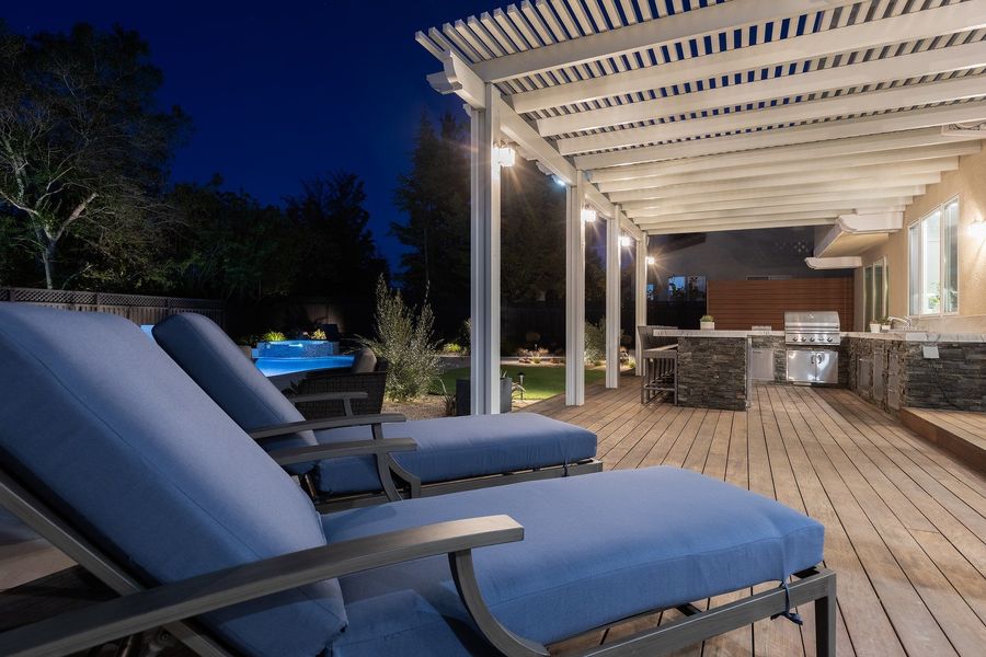 Night view under the pergola past two blue-cushioned loungers along the deck toward the stone-clad kitchen island