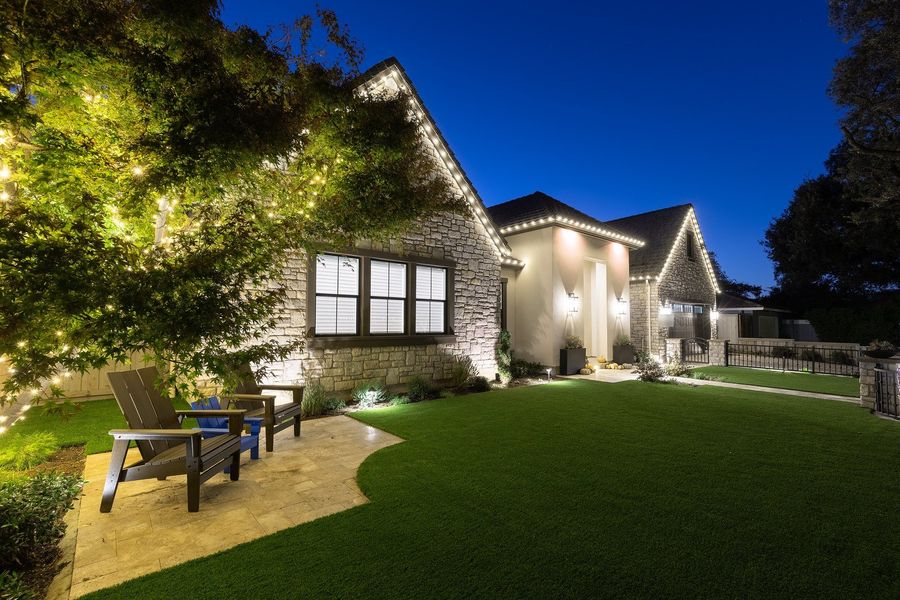 Twilight view across a manicured lawn to a lit stone gable, curved travertine patio with chairs and warm light in the tree