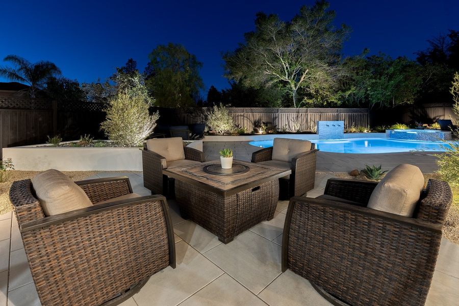 Night view of woven armchairs around a fire table on pale paving, with olive shrubs lit from below and the garden beyond