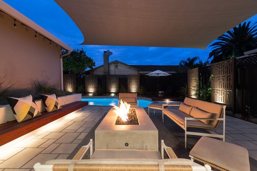 Gas fire table and lit ipe bench with outdoor sofas under a shade sail at blue hour, with uplit decorative screens beyond
