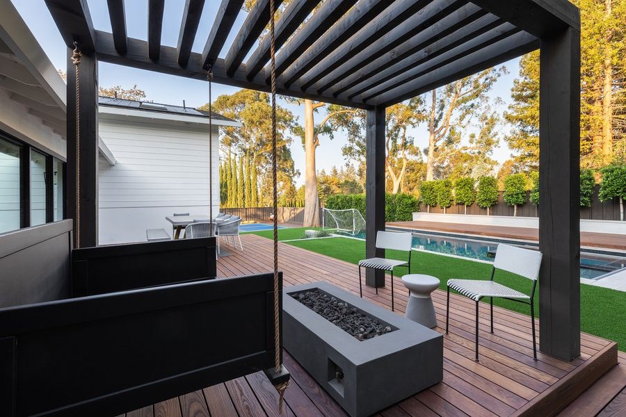 Black timber pergola over a hanging daybed swing and a concrete gas fire table on a hardwood deck with the lawn beyond