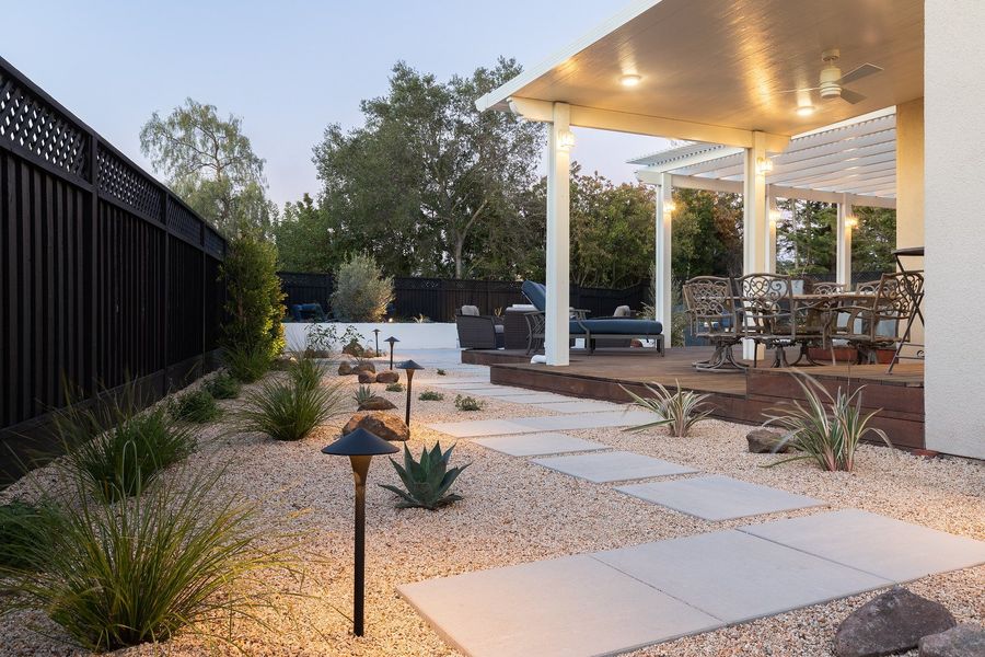 Square pale slabs stepping through a gravel bed of agave and grasses along a dark fence toward a covered patio, with a path light in the foreground