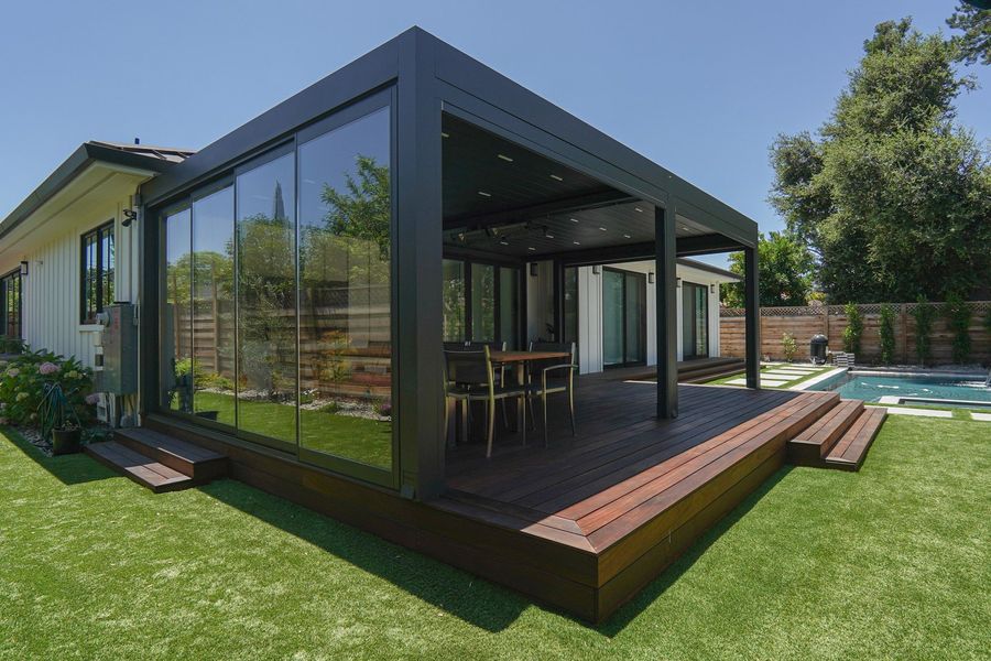 Wide wood deck with low steps in front of a dark steel covered patio with a dining table, and green lawn in front, with a corner of the yard visible at right.