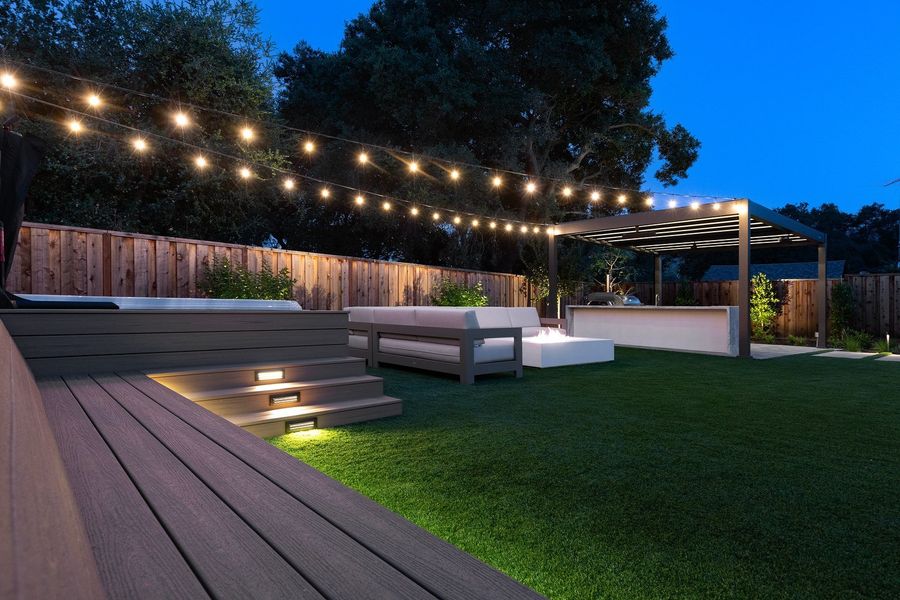 Lit steps on a raised deck beside a sofa and fire table at night, string lights above and a pergola in the distance