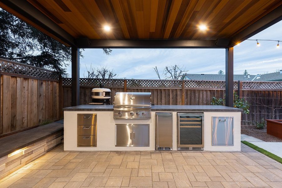 Built-in outdoor kitchen with a stainless grill, pizza oven, drawers, refrigerator and wine fridge under a cedar ceiling with recessed lights