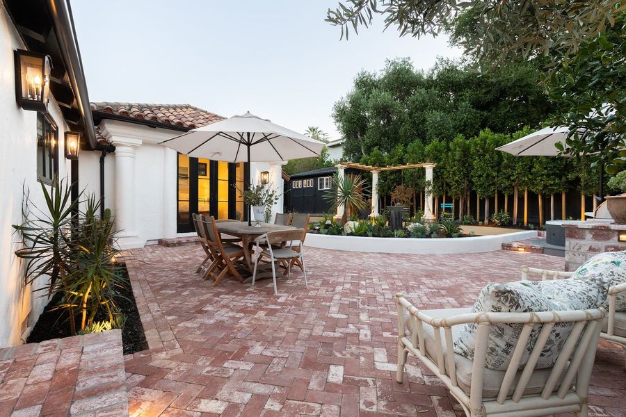 Daytime view across a herringbone brick courtyard with a dining table, a rattan lounge set, a curved planter and a pergola beyond