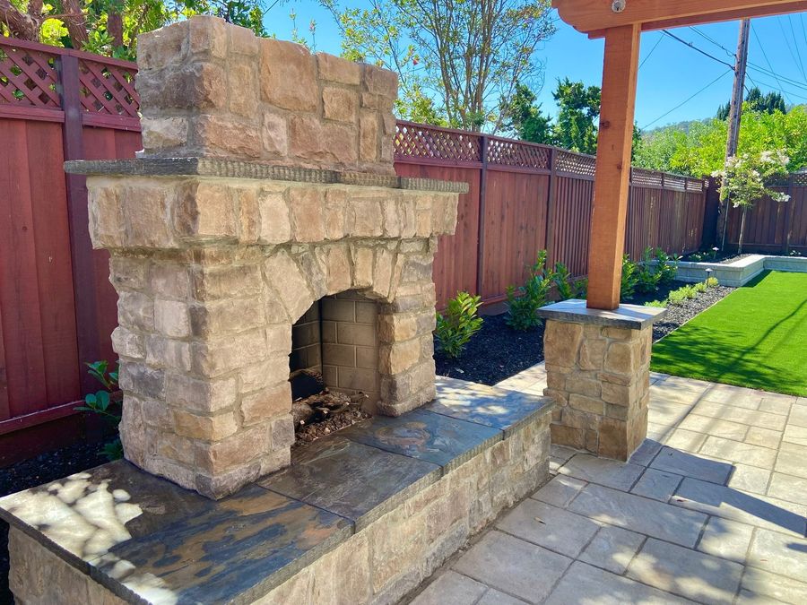 Stacked stone fireplace with an arched opening on a raised stone base with a slate cap, beside a stone pillar and a paver patio.