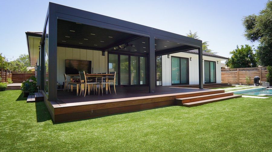 Covered patio with a dining table opening onto a wood deck with steps, bright green lawn in the foreground.