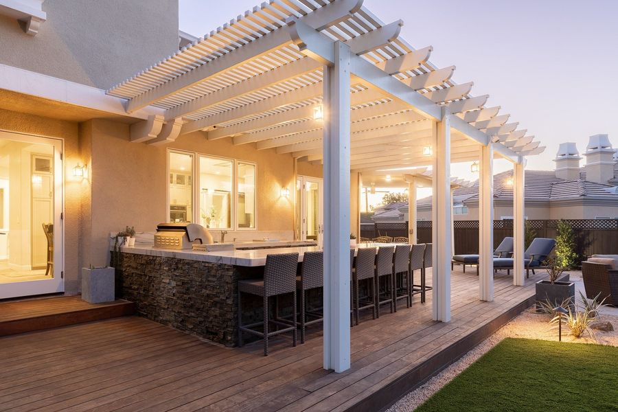 White louvered pergola over a deck with a stone-clad bar counter and tall stools, warm light spilling from the house at dusk