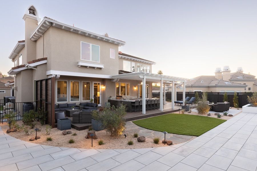 Late-dusk view of the back of the house with a white pergola over the deck, a curved lawn, pale paving slabs and gravel beds with boulders and grasses