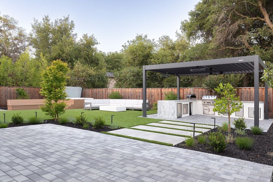 Grey paver terrace with concrete stepping pads set in turf leading toward a louvered pergola and kitchen island at dusk