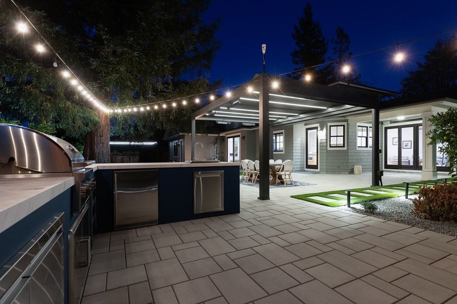 Night view along the stone counter and stainless fridge to the lit pergola and string lights over the terrace
