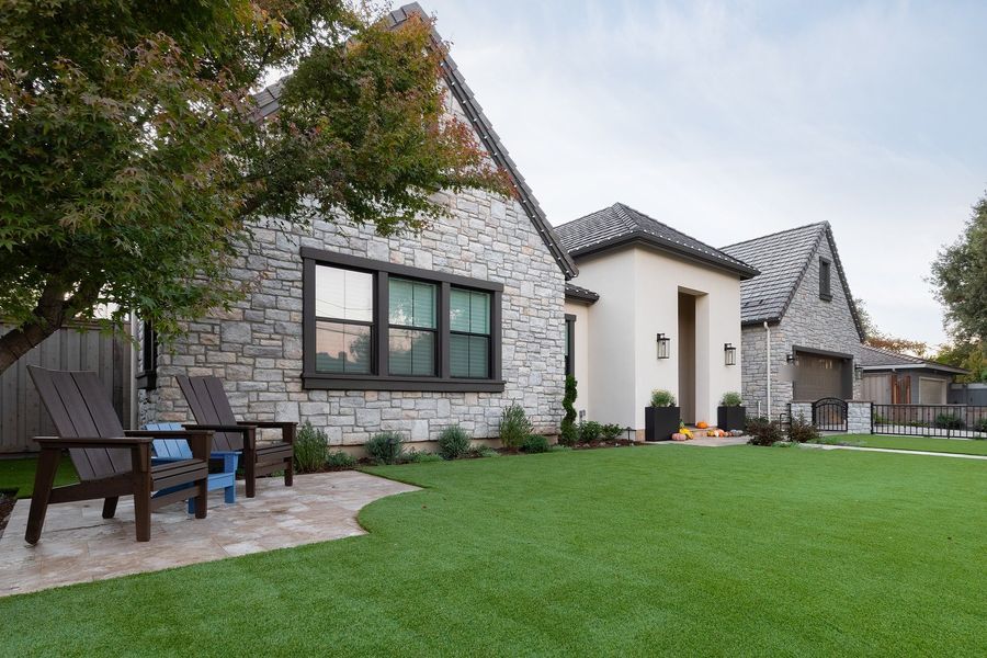 Daytime front garden with green lawn, pale travertine patio with two dark lounge chairs and a stone and stucco house behind
