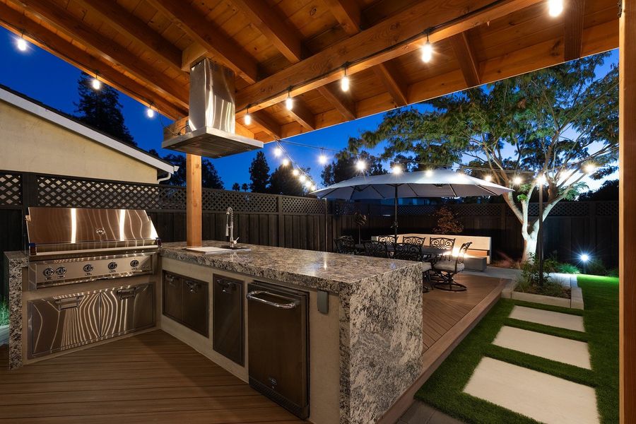 Evening view of a granite-topped outdoor kitchen island with a built-in grill and stainless hood under a timber roof, with string lights and a dining table beyond