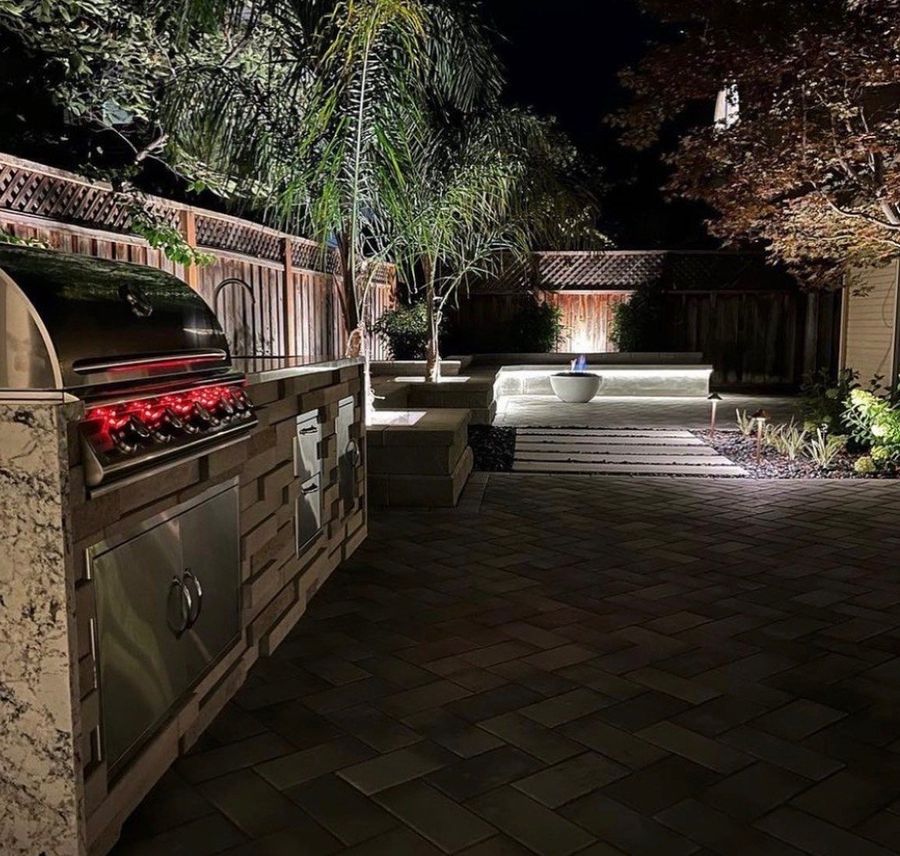 Evening view across the paver patio past the built-in grill with red glowing knobs to a lit seat wall with a white fire bowl showing a small blue flame.