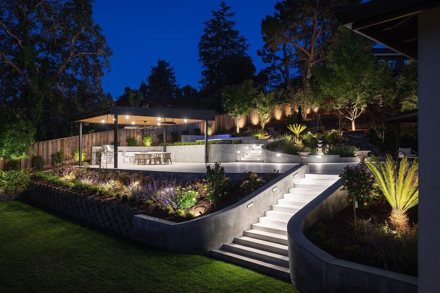 Low night view of the lit stair climbing between curved walls to the glowing pavilion above a lawn