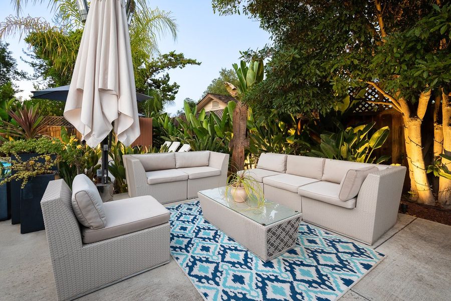 Cream outdoor sectional sofa and a glass coffee table on a blue and white patterned rug, with a folded umbrella and uplit trees and planting at dusk.