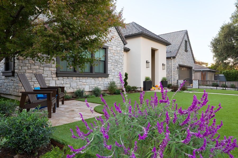 Purple salvia in the foreground with a lawn, travertine patio and stone house with dark window trim