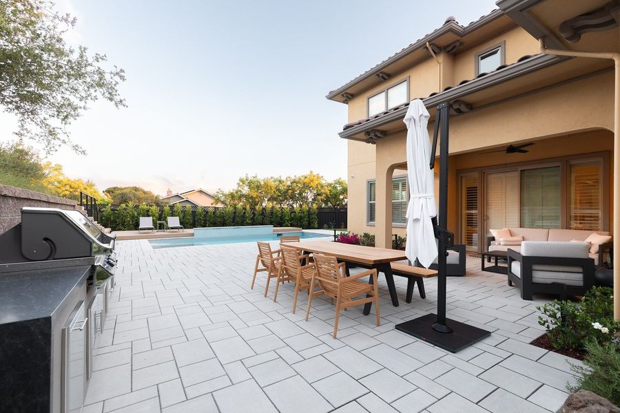 Daytime paver patio with a built-in grill island at left, teak dining table and covered seating area at right