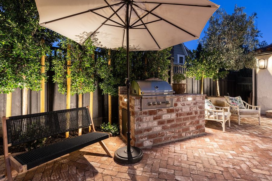Twilight view of a brick grill island with a stainless grill beside a woven bench and an umbrella, with uplit evergreens behind