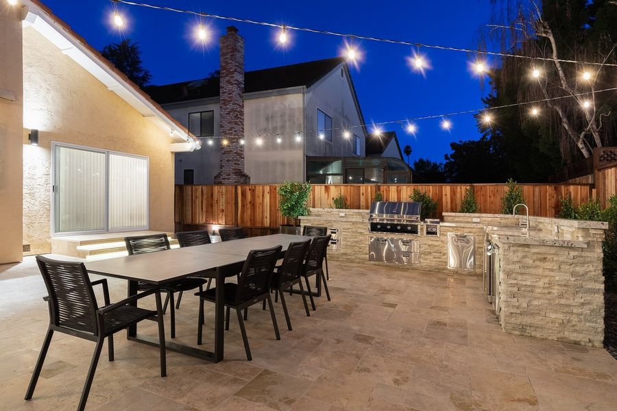 Twilight view of a long dining table with black chairs beside a stacked-stone kitchen island with a built-in grill, under string lights