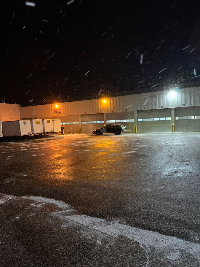 Snow falling at night over a wet parking lot lit by the lights of a long building with trucks parked near it