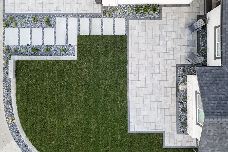 Top-down daytime view of a paver patio, a row of concrete stepping slabs in river rock and a lawn bounded by a curved concrete curb