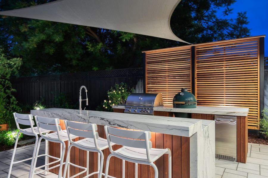 Stone-topped bar with under-counter lighting, four white stools, grill and ceramic cooker in front of a slatted cedar screen at dusk