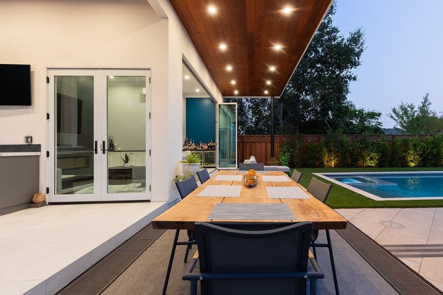 Long timber dining table under the cabana ceiling with recessed lights, glass doors open to the lounge behind