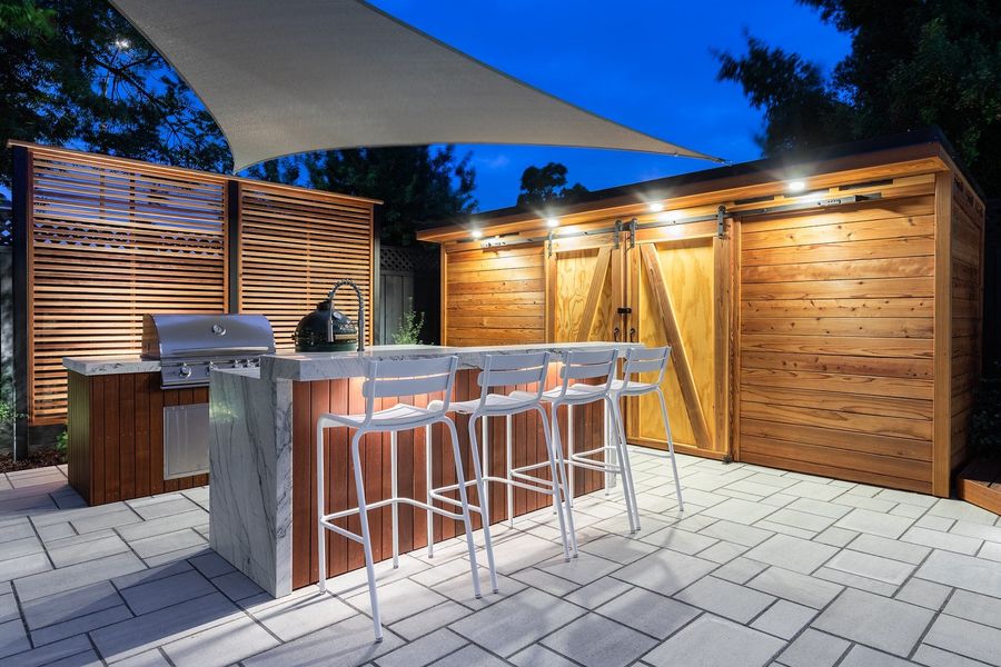 Outdoor kitchen at twilight with built-in grill, waterfall-edge stone bar with white stools and a cedar cabinet with barn doors lit from above