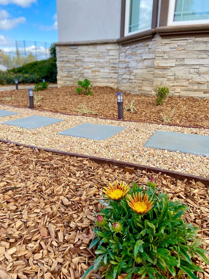 Square gray stone pavers set in pale gravel beside a bed of wood-chip mulch with orange gazania flowers, steel edging and path lights, with stacked stone on the house behind.