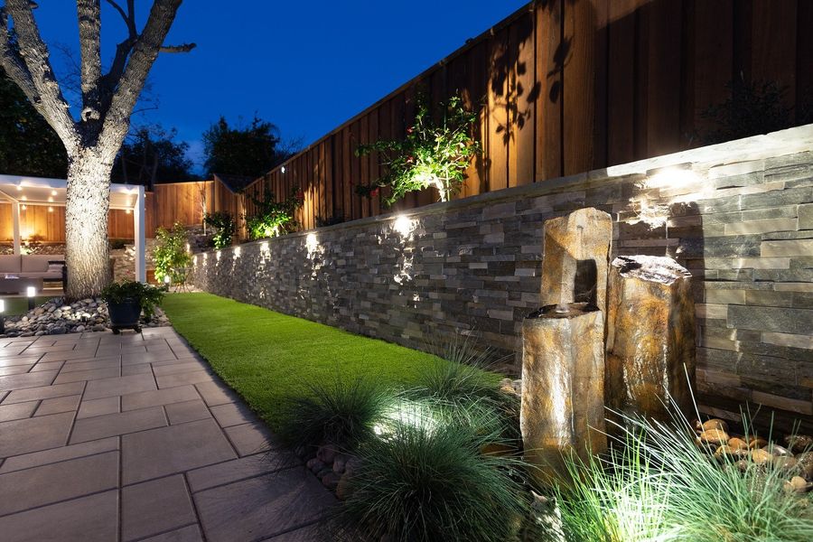 Lit stone wall with a basalt column fountain, blue fescue grass and a paver path running alongside turf at night