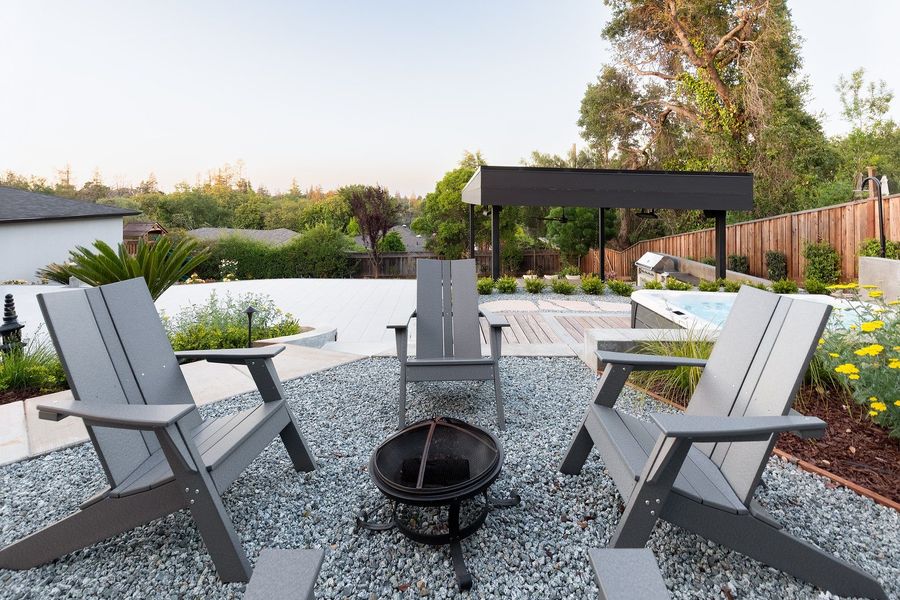 Close view of gray Adirondack chairs and a black fire bowl on pale gravel beside the yellow-flowered planting