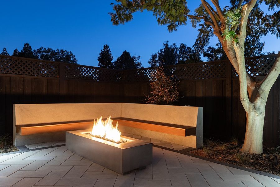 Twilight view of a lit corner bench and a gas fire table on pale pavers beneath a tree