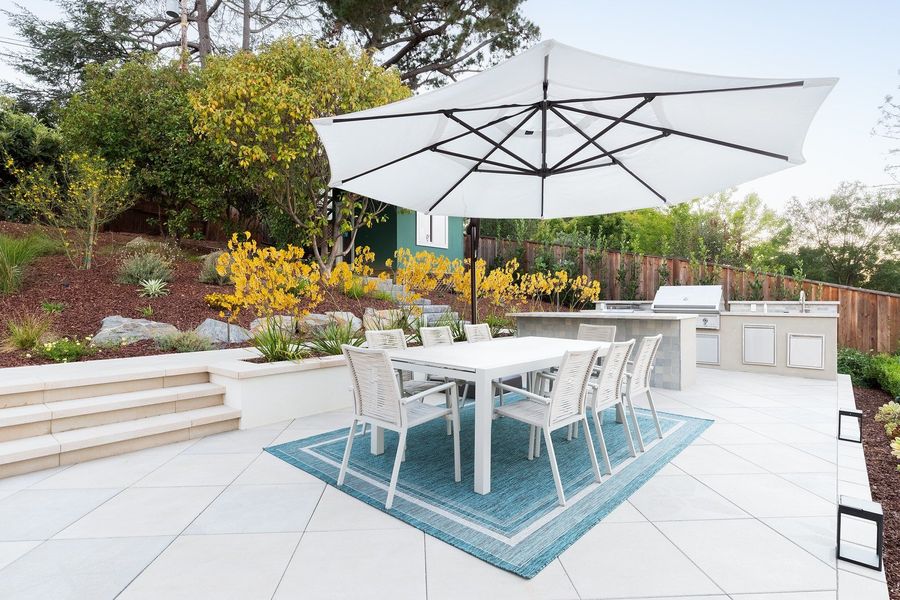 Dining table on a blue rug under a white cantilever umbrella, with the built-in grill island and a small green shed behind