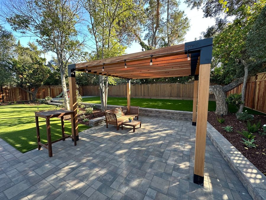 Wood pergola with black steel corners and string lights over a gray paver patio, with a lounge chair, a small table, a bar-height table and a lawn behind.