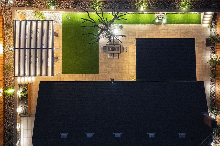 Night aerial of the backyard with a turf strip, a dining set on paving, a white louvered pergola in the corner and lights along the walls