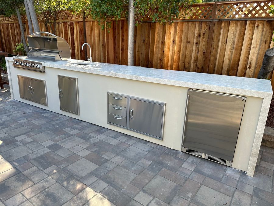 Long white stucco outdoor kitchen island with a stainless grill, sink, access doors, drawers and refrigerator under a pale stone countertop, on gray pavers.
