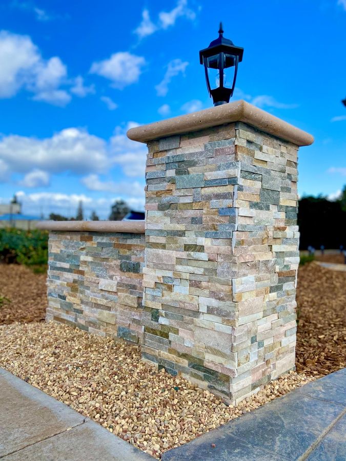 Stacked multicolor stone pillar with a cream cap and a black lantern on top, a low stone wall and pale gravel, against a blue sky with clouds.