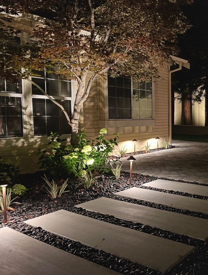 Evening view of pale stepping-stone slabs in black pebbles with copper path lights, glowing hydrangeas and variegated grasses, and a lit paver path along the house.