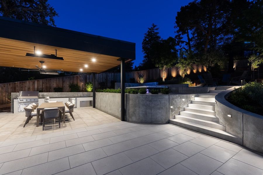 Night view of the lit concrete stair and curved wall beside the glowing pavilion kitchen with a long dining table