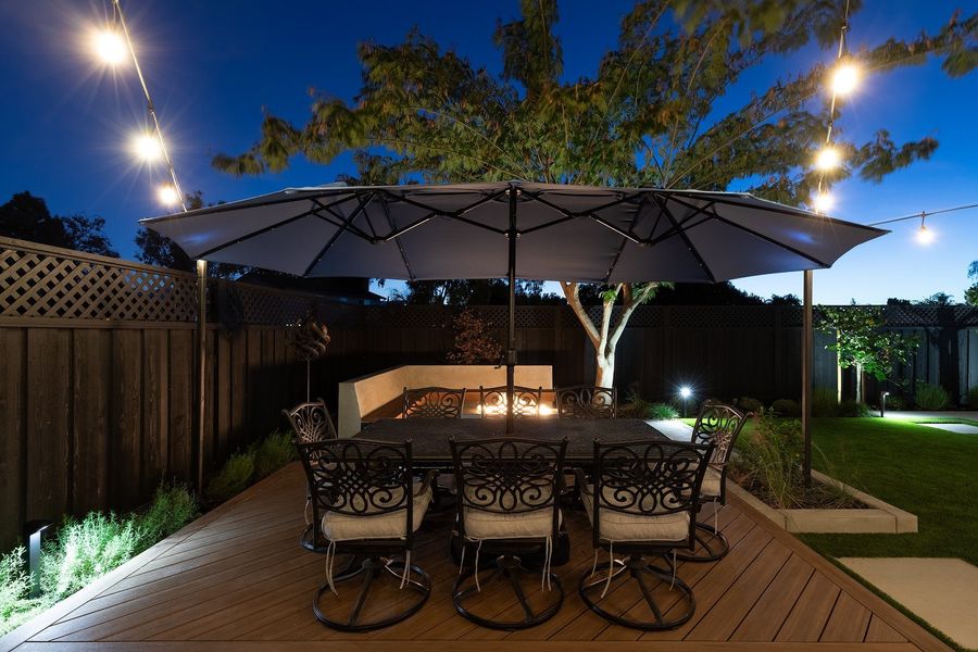Evening view of a dining table under a large umbrella on a wood-look deck, with string lights, uplit planting and a tree overhead