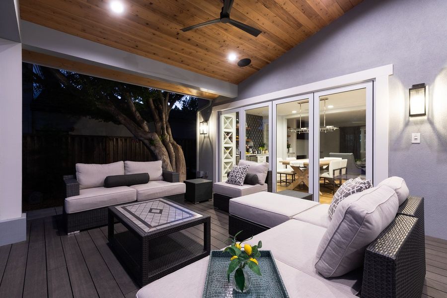 Night view of woven sofas on the deck beneath a vaulted cedar ceiling, with a mature tree trunk beyond the open side and a glowing wall lantern
