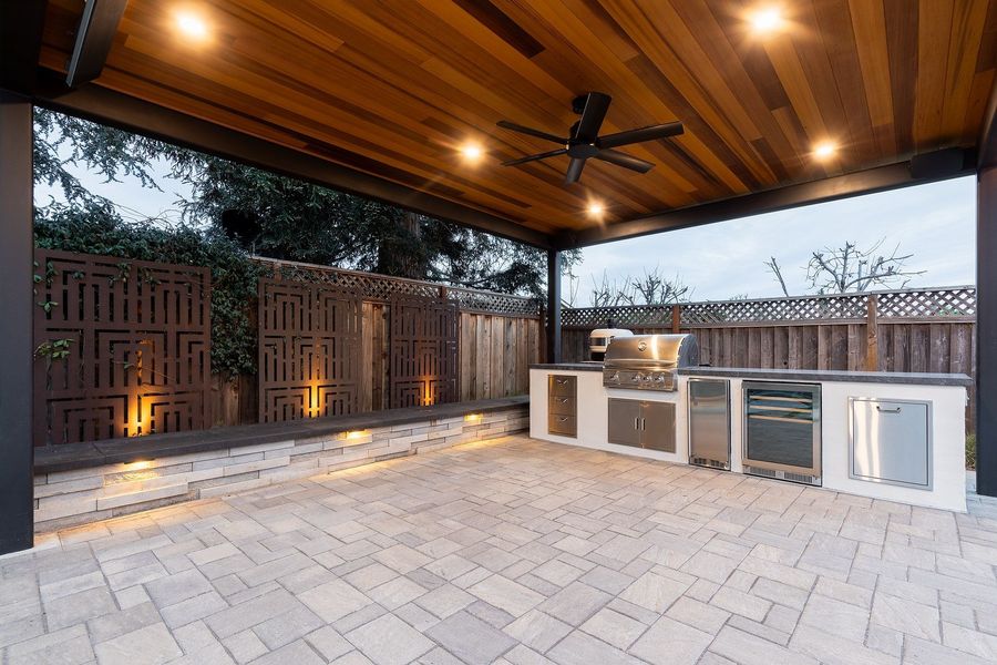 Kitchen under the cedar ceiling at dusk with uplit patterned metal screens and a lit stacked-stone seat wall