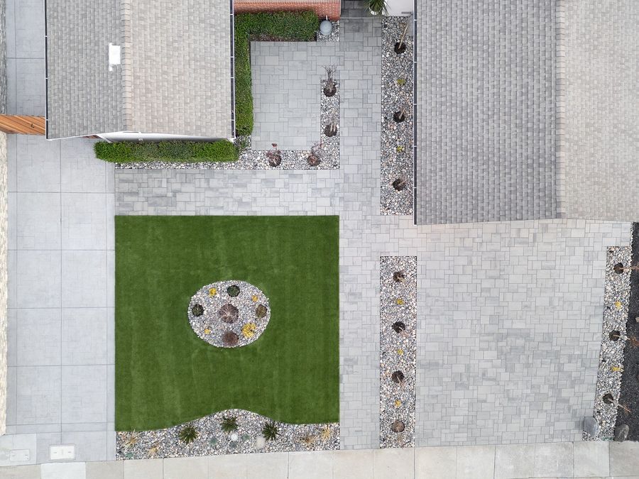 Aerial view of a square of turf with a round bed of stones and plants, framed by paver driveway, paths and river-rock strips with young trees