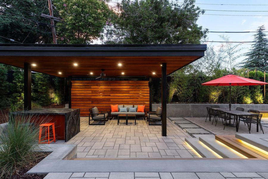 Flat-roofed pavilion with a wood ceiling and recessed lights, a lounge set, a stone-topped bar and lit steps beside the dining terrace