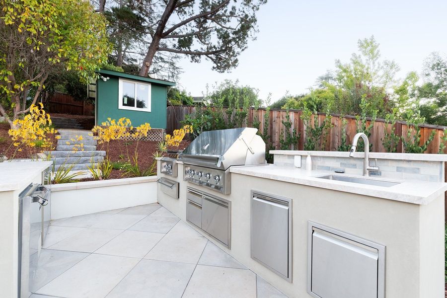 Stucco outdoor kitchen island with a stainless grill, sink, stone worktop and stainless access doors on pale paving