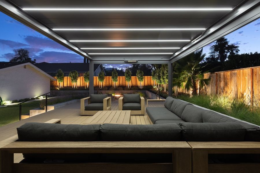 Night view from inside the pergola with white strip lights overhead, dark sofa and a glowing hedge line beyond
