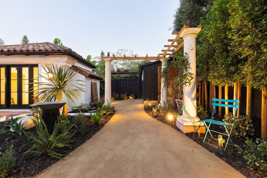 Decomposed granite path winding through agave and fern planting toward a column pergola, with the lit back doors at left
