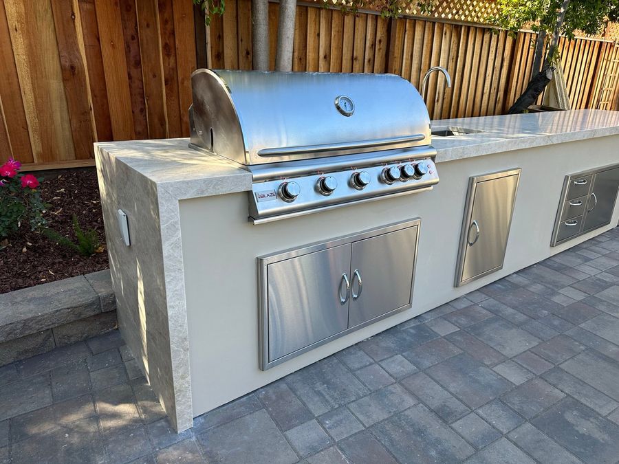 Close view of a stainless steel built-in grill and double access doors in a white stucco island with a pale stone countertop edge, on gray pavers.